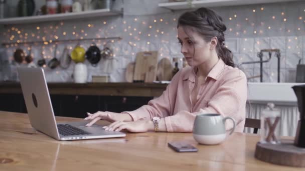 Cute brunette woman looking deep in thought while sitting at a table in her kitchen, working online with a laptop, freelance concept. Stock footage. Young woman using her device. — ストック動画