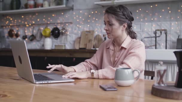 Cute brunette woman looking deep in thought while sitting at a table in her kitchen, working online with a laptop, freelance concept. Stock footage. Young woman using her device. — ストック動画