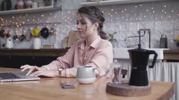 Una joven sentada en la mesa de la cocina usando un portátil. Imágenes de archivo. Hermosa morena trabajando en casa en su ordenador con una taza de bebida caliente de pie sobre la mesa de madera . — Vídeo de stock