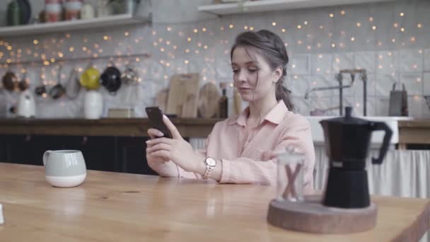 Young woman sitting at her kitchen table at home working on her small business with a smart phone. Stock footage. Portrait of a young lady tapping oh her mobile phone. — Stock Video