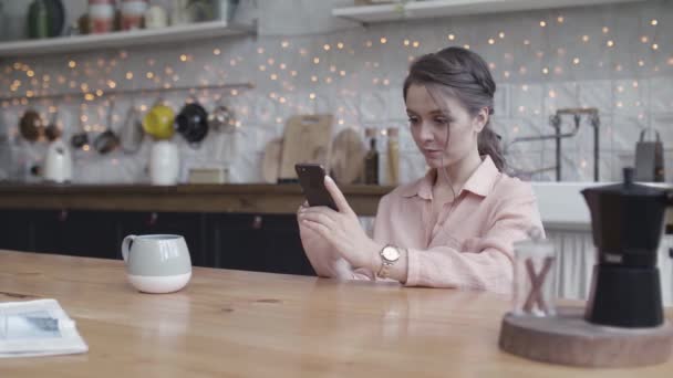 Een jonge vrouw met haar mobiele telefoon en een kop koffie in de keuken, communicatieconcept. Voorraadbeelden. Jonge mooie dame afluisteren en sms 'en op haar smartphone thuis. — Stockvideo