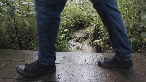 Közelkép a turisták lábait séta a fa hídon a piszkos erdei patak felett. Készletfelvétel. Oldalnézet túrázók átkelés a fa kis híd felett keskeny folyó. — Stock Fotó