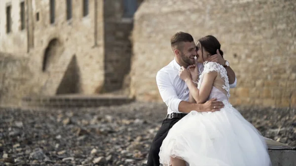 Beautiful fairytale newlywed couple sitting together and hugging, sharing love on high brick wall baackground. Action. Groom and bride during photosession near old medieval castle — Stock Photo, Image