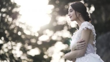 Bulanık yeşil çalıların ve ağaçların arka planında beyaz gelinlik giymiş bir gelinin portresi. Başla. Şehvetli kahverengi saçlı kız yan bakıyor, gülümsüyor ve kendine sarılıyor, düğün konsepti..