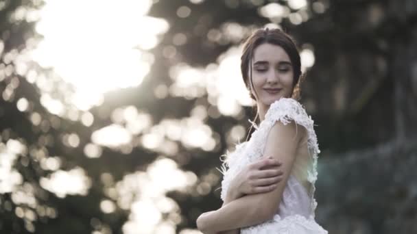 Portrait of a bride in a white wedding dress on a background of blurred green bushes and trees. Action. Sensual brown-haired girl looking aside, smiling and hugging herself, wedding concept. — ストック動画