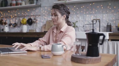 Mutfak masasında dizüstü bilgisayar kullanan genç bir kadın. Stok görüntüleri. Evinde bilgisayarında çalışan güzel bir esmer ve bir bardak sıcak içecekle ahşap masada duruyor..