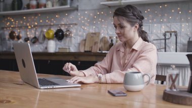 Şirin esmer kadın mutfağındaki bir masada oturmuş dizüstü bilgisayarla internetten iş yaparken derin düşüncelere dalmış. Stok görüntüleri. Genç kadın aletini kullanıyor..