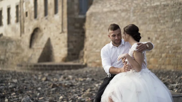 Fotosessione all'aperto di amare gli sposi alla moda sulla riva pietrosa vicino al castello. Azione. Bella coppia di nozze che abbraccia e sorride, concetto di famiglia e amore . — Foto Stock