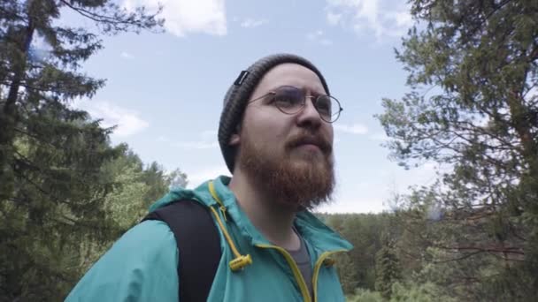 Close up of hiker man looking aside and then passing by the camera in the forest. Stock footge. Bearded man in round glasses walking through the woods, tourism concept. — ストック動画