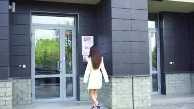 Uzun siyah saçlı, beyaz önlüklü ve yüksek topuklu sandaletli genç ve güzel bir kadının arka görüntüsü ticari bir binaya girmek üzere. - Sanat. Kadın kapıyı açıyor ve binaya giriyor..