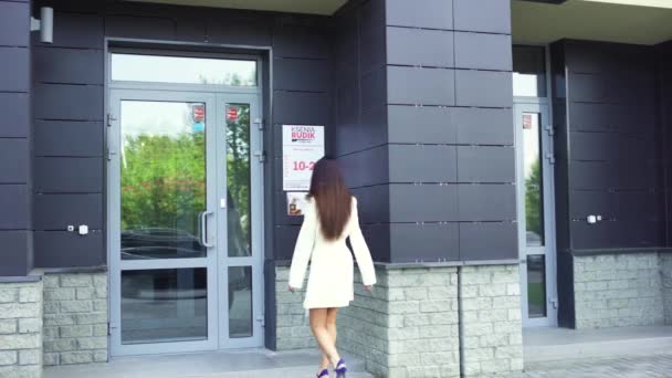 Visão traseira de uma mulher jovem e bonita com cabelo escuro longo em casaco branco e sandálias de salto alto está prestes a entrar em um edifício comercial. Arte. Mulher abrindo a porta e entrando no prédio . — Vídeo de Stock