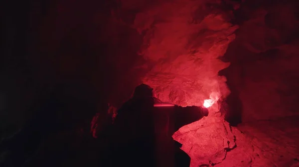 Viajero con llamarada de señal roja en la cueva oscura, vista inferior. Imágenes de archivo. Joven explorando la oscura cueva brumosa y dejando atrás la entrada . — Foto de Stock