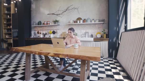 Mujer atractiva joven que trabaja en el ordenador portátil en casa. Imágenes de archivo. Hermosa joven mujer atentamente trabajando en el ordenador portátil sentado en la mesa en la cocina con estilo — Vídeos de Stock