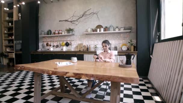 Jovem mulher atraente sentado no telefone na mesa da cozinha. Imagens de stock. Jovem descansa durante o almoço com telefone e xícara de café na cozinha — Vídeo de Stock