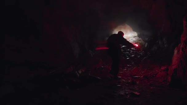 Silhouette of a cave explorer in the underground with the entrance behind him, tourism and nature concept. Stock footage. Male tourist walking inside the cave with burning signal flare. — Stock Video