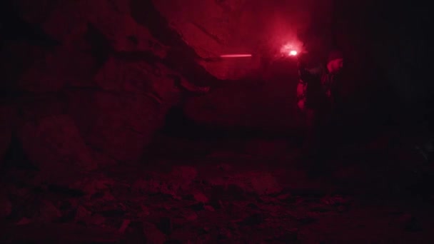 Sylwetka człowieka z czerwonym palącym się sygnałem rozbłysku chodzącego po kamieniach w ciemności jaskini, ekstremalna koncepcja turystyki. Materiał filmowy. Mężczyzna odkrywca wewnątrz podziemnego tunelu. — Wideo stockowe