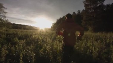 Uzun çimenlerle vahşi yeşil arazide koşan bir adamın arka görüntüsü. Stok görüntüleri. Kamera koşucuyu güzel bir sonbaharda, günbatımı çayır arka planında takip eder.