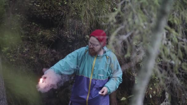 El joven sostiene una llamarada de humo rojo para pedir ayuda en medio de la naturaleza salvaje y verde. Imágenes de archivo. Hipster viajero perdido en el bosque y pidiendo ayuda, SOS . — Vídeos de Stock