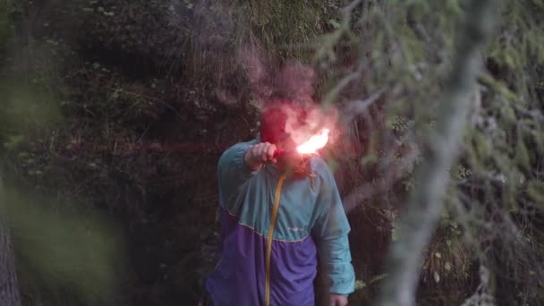 Hombre barbudo con chaqueta colorida y gafas redondas balanceándose llamarada de señal roja ardiente y esperando a ser encontrado. Imágenes de archivo. Turista masculino perdido en el bosque verde . — Vídeos de Stock