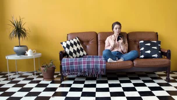 Belle jeune femme assise sur un canapé avec téléphone. Images d'archives. Femme attrayante retournant par fil d'actualité sur le téléphone assis dans le salon confortable — Video