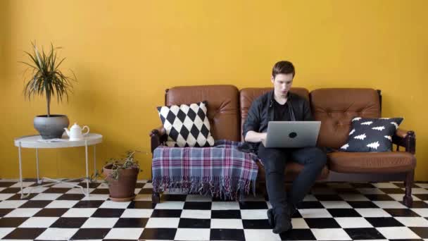 El joven trabaja en el portátil sentado en el sofá. Imágenes de archivo. Joven trabaja en el modo independiente en el portátil, sentado en el sofá en el interior del diseñador. Joven freelancer está trabajando intensamente en el portátil — Vídeo de stock