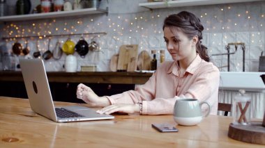 Genç ve çekici bir kadın evde, mutfakta dizüstü bilgisayarla çalışıyor. Stok görüntüleri. Güzel bir kadın dizüstü bilgisayarda serbest modda ve rahat mutfağın arka planında dikkatlice çalışıyor.