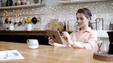 Genç ve çekici bir kadın, rahat bir mutfakta tablette çalışıyor. Stok görüntüleri. Genç ve şık bir kadın evdeki tablette tasarım projeleri yapıyor. Rahat evdeki tablette serbest çalışma