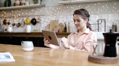 Genç ve çekici bir kadın, rahat bir mutfakta tablette çalışıyor. Stok görüntüleri. Genç ve şık bir kadın evdeki tablette tasarım projeleri yapıyor. Rahat evdeki tablette serbest çalışma