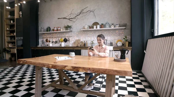 Młoda, atrakcyjna kobieta siedząca przy stole kuchennym. Materiał filmowy. Młoda kobieta odpoczywa podczas lunchu z telefonem i filiżanką kawy w kuchni — Zdjęcie stockowe
