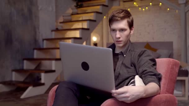 Thuis werken. Voorraadbeelden. Vrolijke jonge knappe man werkt op laptop in grote comfortabele stoel thuis — Stockvideo