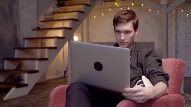 这个年轻人坐在沙发上，在笔记本电脑上工作。 库存录像。 年轻的自由职业者正在专心致志地工作在笔记本电脑上 — 图库视频影像