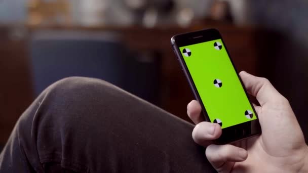 Man met mobiele smartphone thuis. Voorraadbeelden. Close-up van een telefoon met een groen scherm in de handen van een man — Stockvideo