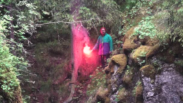 Uomo escursionista in abiti luminosi osservando le piante verdi della scogliera e tenendo un bagliore di segnale rosso in fiamme. Filmati delle scorte. Explorer perso nella foresta, concetto SOS . — Video Stock