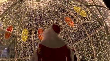 Noel Baba 'nın inşaatın altında ışıltılı ışıklarla yürüdüğünü görmek, Mutlu Noeller ve yeni yıl konsepti. Şarjör. Noel Baba çelenklerin altında.