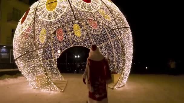 Rear view of Santa Clous coming to the Christmas lights decorations in the street at night, winter holidays concept. Clip. Amazing spherical construstion covered with garlands. — 비디오