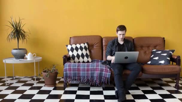 Een jongeman die thuis aan een laptop werkt. Voorraadbeelden. Hij werkt thuis als freelancer op een laptop op de bank. Moderne werkstijl thuis in freelance modus — Stockvideo