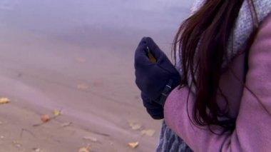 Elinde sarı bir yaprak tutan bir kadın kumlu bir kıyıda ve nehirde. Stok görüntüleri. Pembe ceketli, koyu mavi eldivenli, sarı bir ağaç yaprağını tutan bir kadının fotoğrafını çek..