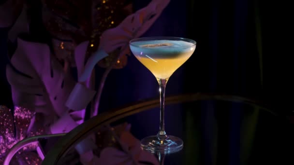 Erfrischendes alkoholisches Getränk im Martini-Glas. Archivmaterial. Großaufnahme eines Getränks, das auf einem Glastisch auf schwarzem Hintergrund mit fliederfarbener Kunstpflanze serviert wird. — Stockvideo