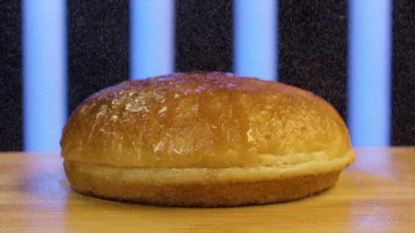 Close up of hamburger bun cut in two. Stock footage. Wheat burger bun lying on wooden surface with flying flour on black background with blue stripes. — Stock Photo, Image