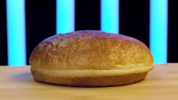 Close up of hamburger bun cut in two. Stock footage. Wheat burger bun lying on wooden surface with flying flour on black background with blue stripes. — Stock Video