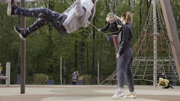 Retrato de una pequeña hija y madre jugando swing en el parque mientras sonríen juntas. Imágenes de archivo. Madre empujando a su hija en un columpio en el patio al aire libre, concepto familiar . — Foto de Stock