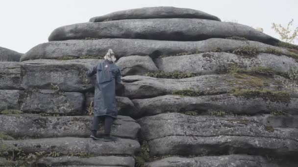 Vue arrière d'un homme vêtu d'une longue veste et aux cheveux colorés essayant d'escalader une montagne pierreuse. Images d'archives. Homme randonneur escalade rochers géants sur fond de ciel nuageux . — Video