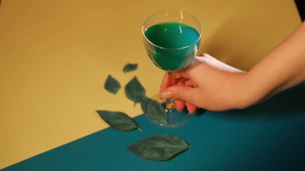 緑の葉でカラフルな表面にブルーアルコールカクテル。ストック映像だ。人工的な葉と黄色と青の生地から珍しい飲料とガラスを取って女性の手の閉じると — ストック動画