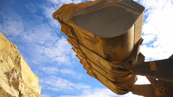 Grävmaskin i drift på byggarbetsplatsen. Lagerbilder. Botten syn på grävmaskin hink med gul färg laddad med sand på blå molnig himmel bakgrund. — Stockvideo