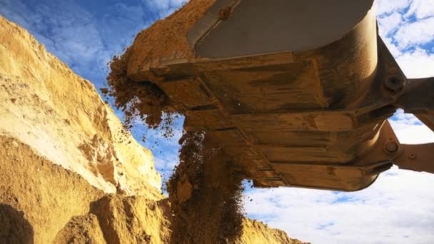 Escavadeira em ação no canteiro de obras. Imagens de stock. Vista inferior da escavadeira lançando o balde de cor amarela carregado com areia no fundo azul do céu nublado . — Vídeo de Stock