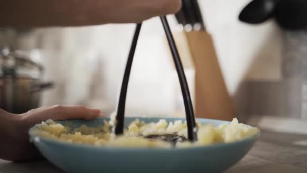 Fechar o processo de fazer purê de batatas. Acção. Mulher mãos fazendo purê de batatas com um espremedor de plástico preto em uma placa azul de vidro na cozinha . — Vídeo de Stock