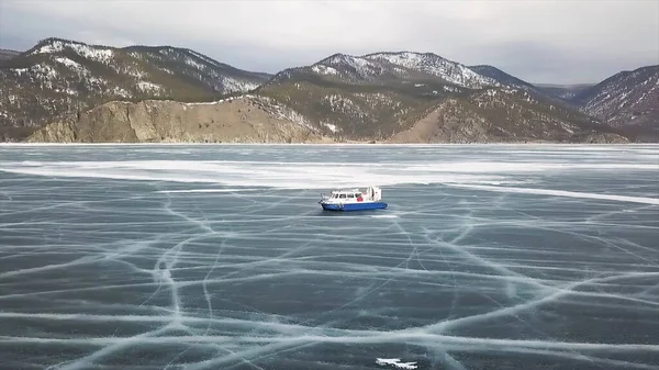 Paseos en aerodeslizador en el lago Baikal. Clip. Vista aérea del vehículo de cojín de aire deslizándose sobre un hermoso lago congelado con montañas cubiertas de nieve, boscosas y cielo nublado en el fondo . — Foto de Stock