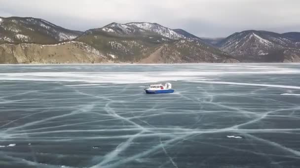 Hovercraft rides on lake Baikal. Clip. Aerial view of air cushion vehicle gliding on beautiful frozen lake with snow covered, forested mountains and cloudy sky on the background. — Stock Video