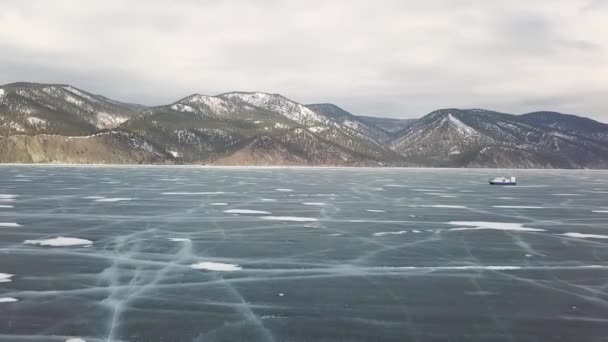 L'aéroglisseur Khivus roule sur le lac Baïkal gelé. Clip. Vue aérienne d'un aéroglisseur se déplaçant sur la glace d'un réservoir gelé en hiver sur des collines enneigées à couper le souffle et un ciel nuageux. — Video