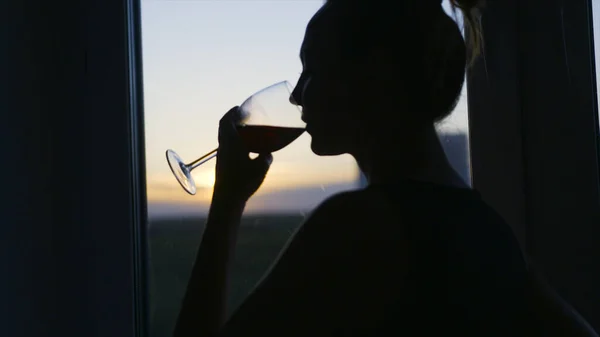 La donna beve il vino con piacere su sfondo di finestra. Azione. Attraente donna beve vino rosso a casa. Silhouette di donna che beve vino sullo sfondo della finestra serale — Foto Stock